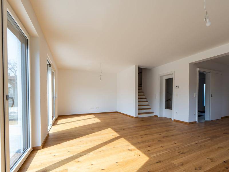 Helles, leeres Wohnzimmer mit Eichenparkett, großen Fenstern und Zugang zur Treppe.