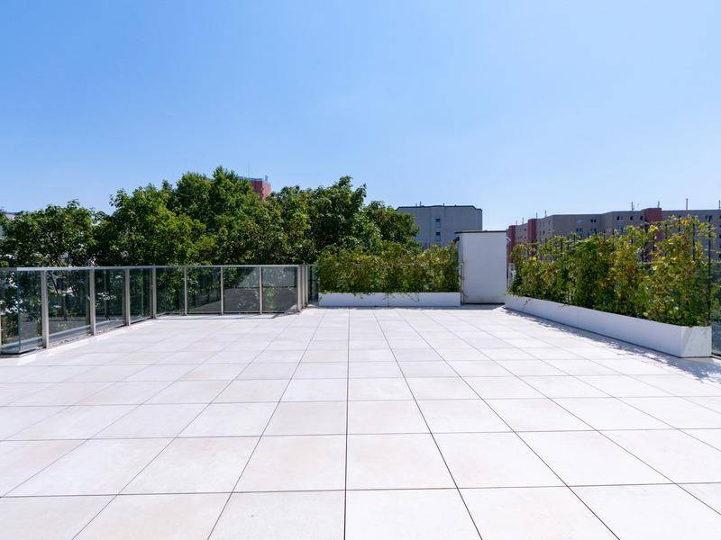 Geräumige Dachterrasse mit Fliesenboden, grünen Pflanzkübeln und Glasgeländer mit Stadtblick.