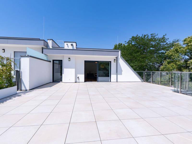Weitläufige, geflieste Dachterrasse mit moderner Architektur und Glasgeländer unter blauem Himmel.