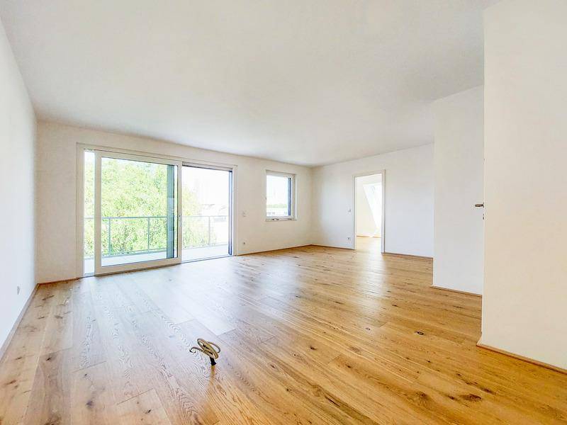 Geräumiges, leeres Wohnzimmer mit hellem Parkettboden und großem Schiebetür zum Balkon.