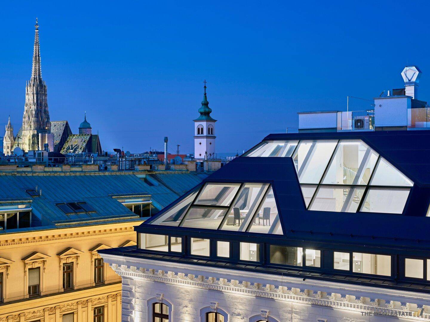 Historisches Gebäude mit modernem Dachausbau und Blick auf die beleuchtete Stadt bei Dämmerung.