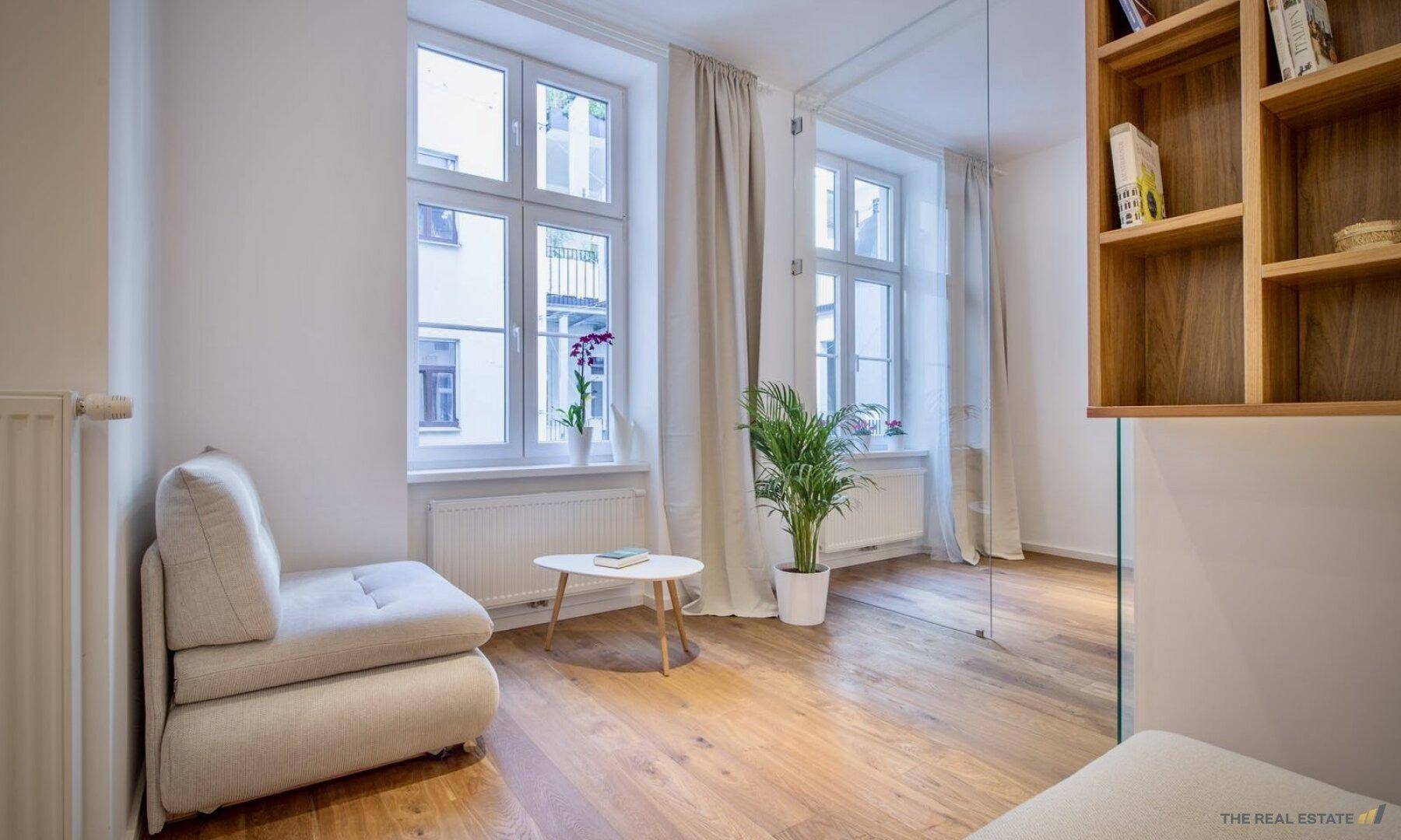 Heller Wohnbereich mit Holzboden, großen Fenstern und einem Bücherregal.