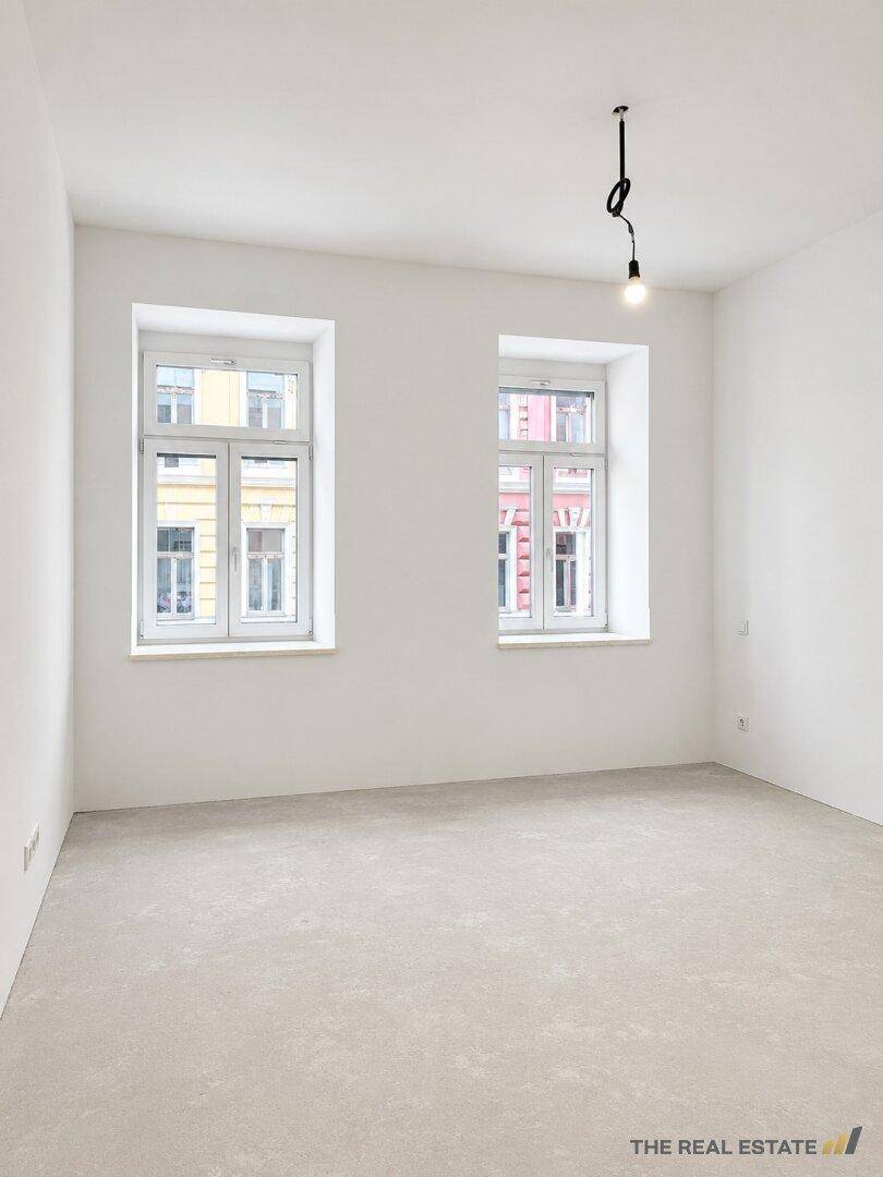 Leerer Raum mit zwei klassischen Fenstern, weißen Wänden und Betonboden.