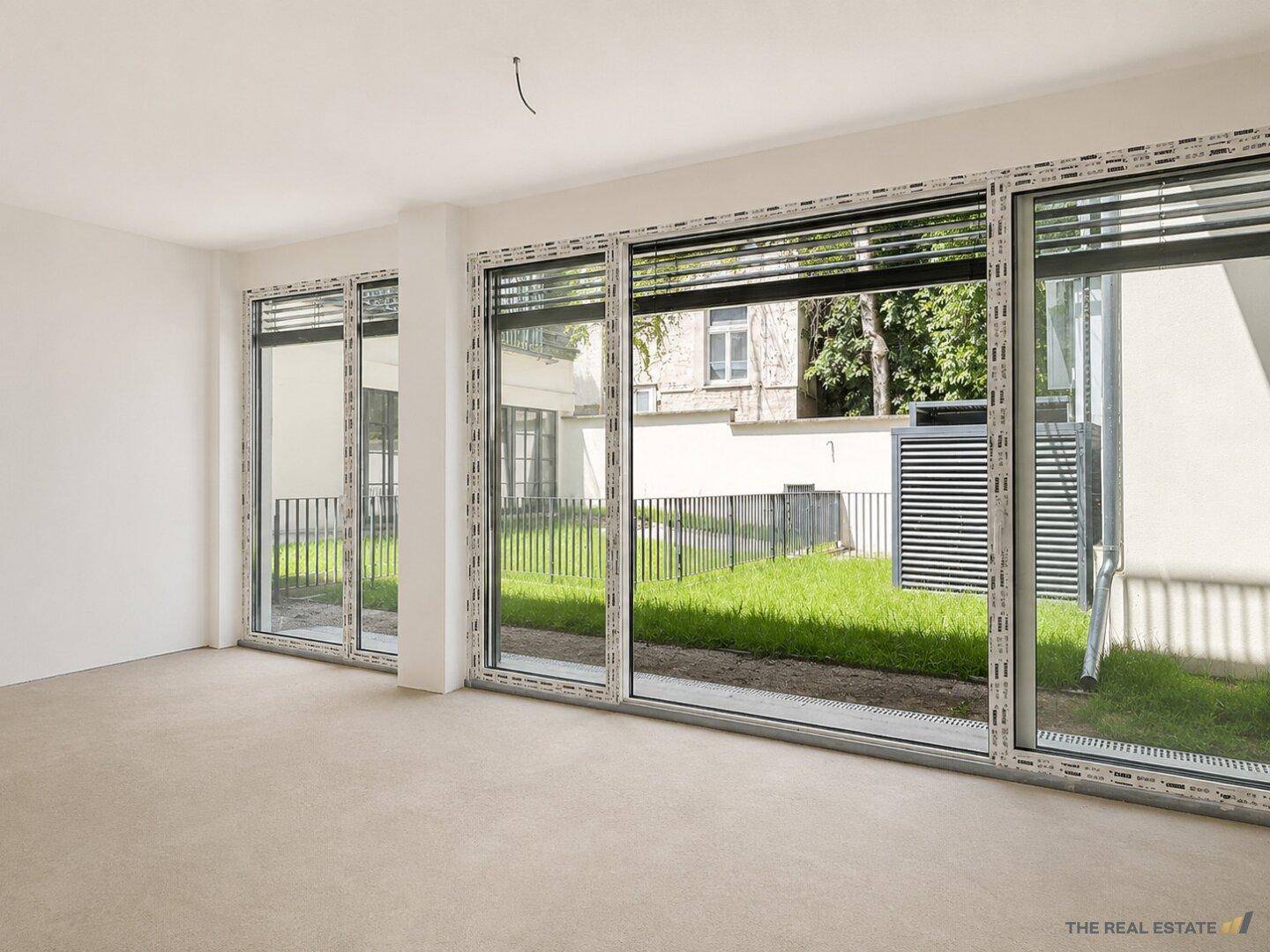 Leerer Raum mit großen Fenstern und Blick auf einen kleinen begrünten Innenhof.