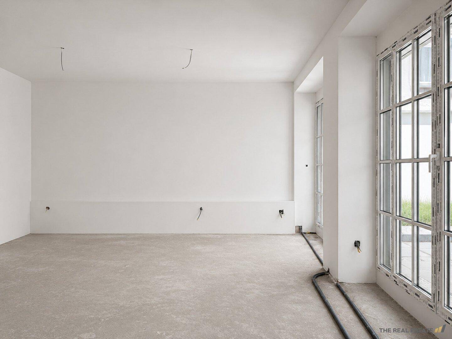 Unmöblierter Raum im Rohbauzustand mit großen Fenstern und Betonboden.