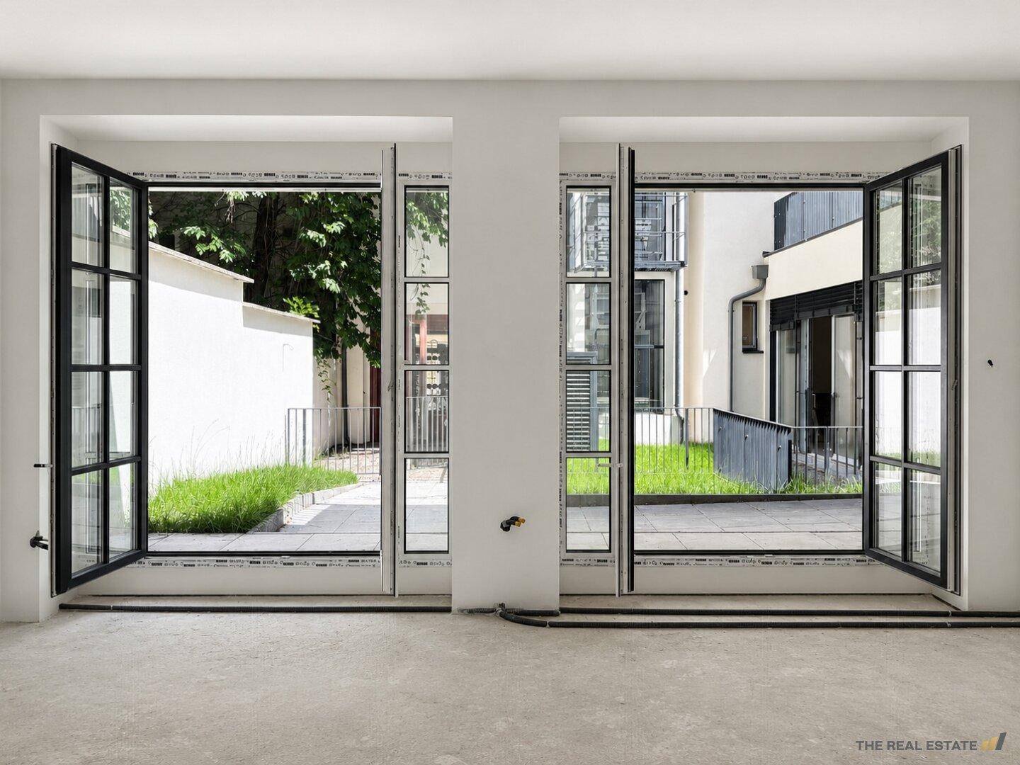 Heller Raum mit geöffneten Fenstertüren und Zugang zu einem begrünten Außenbereich.