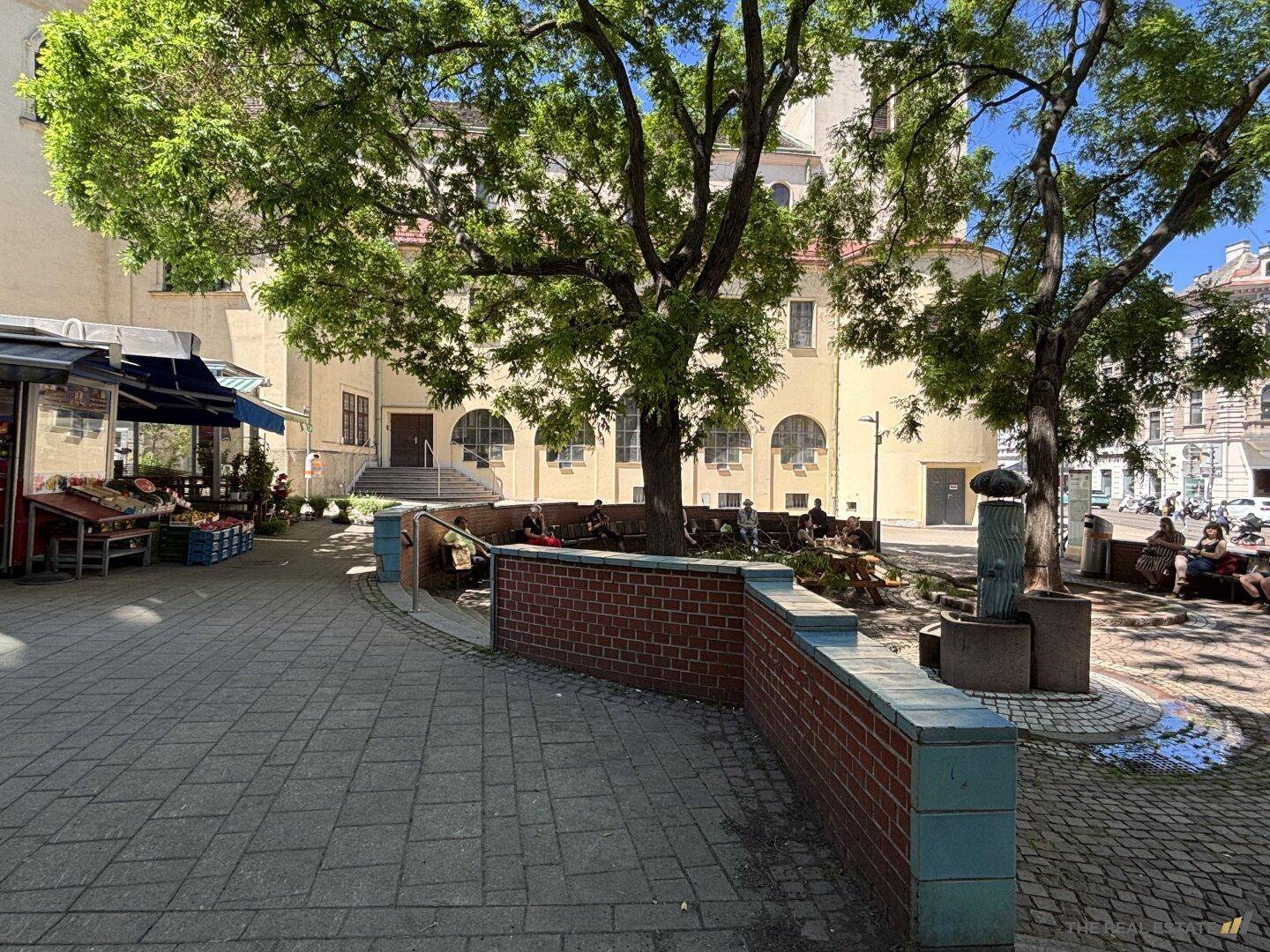Öffentlicher Platz mit Bäumen, Sitzgelegenheiten und einem Brunnen vor historischen Gebäuden.