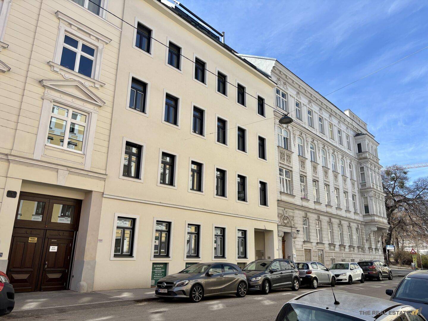 Straßenansicht mit mehrstöckigen Wohngebäuden und geparkten Autos unter blauem Himmel.