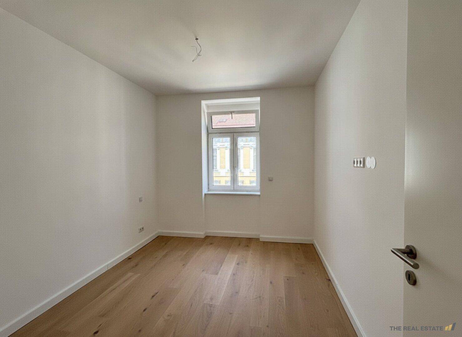 Helles, leeres Zimmer mit Holzboden und einem Fenster mit Blick nach draußen.