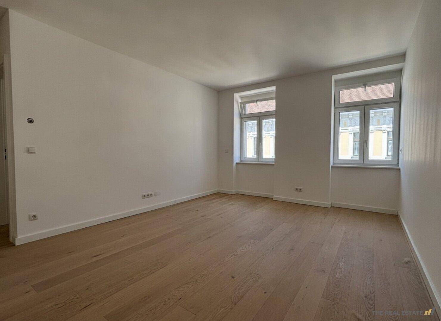 Geräumiges, leeres Zimmer mit zwei Fenstern, Holzboden und viel Tageslicht.