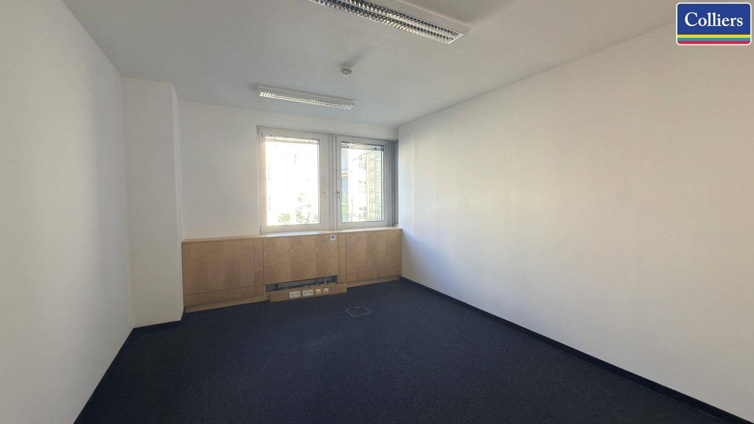Schlichter Büroraum mit weiß gestrichenen Wänden und dunklem Teppichboden, bereit für die Einrichtung eines Arbeitsplatzes.