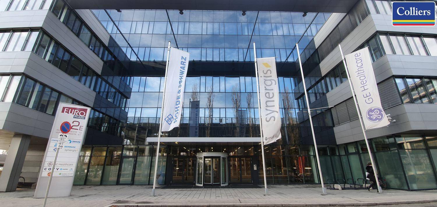 Eingangsbereich eines modernen Bürogebäudes mit großer Glasfassade und mehreren Fahnen davor.