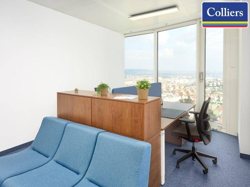Helles Büro mit Schreibtisch, Bürostuhl und einem blauen Sofa, das einen weiten Blick über die Stadt ermöglicht.