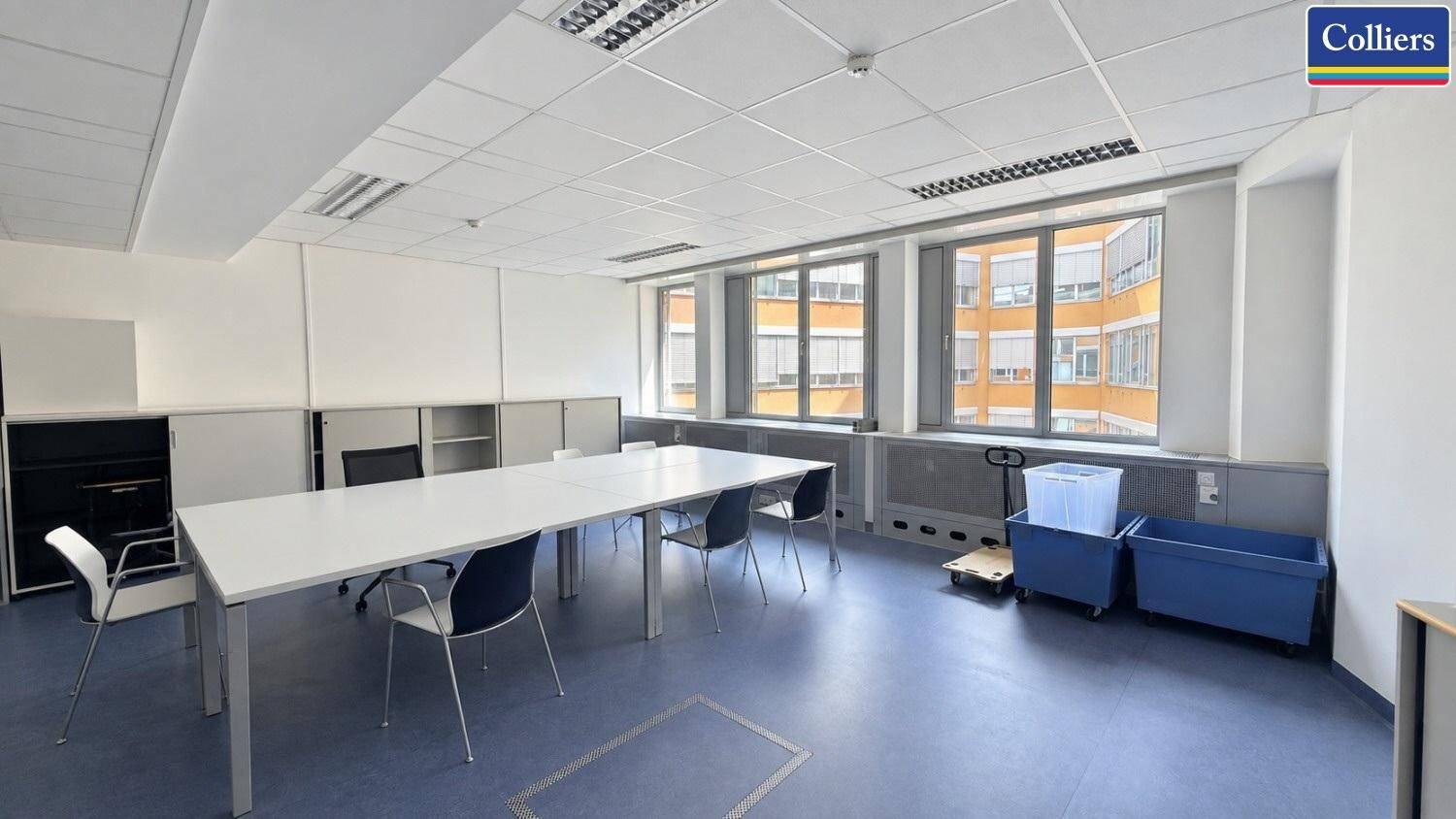 Helles Büro mit langen Tischen, blauem Boden und Blick auf umliegende Gebäude.