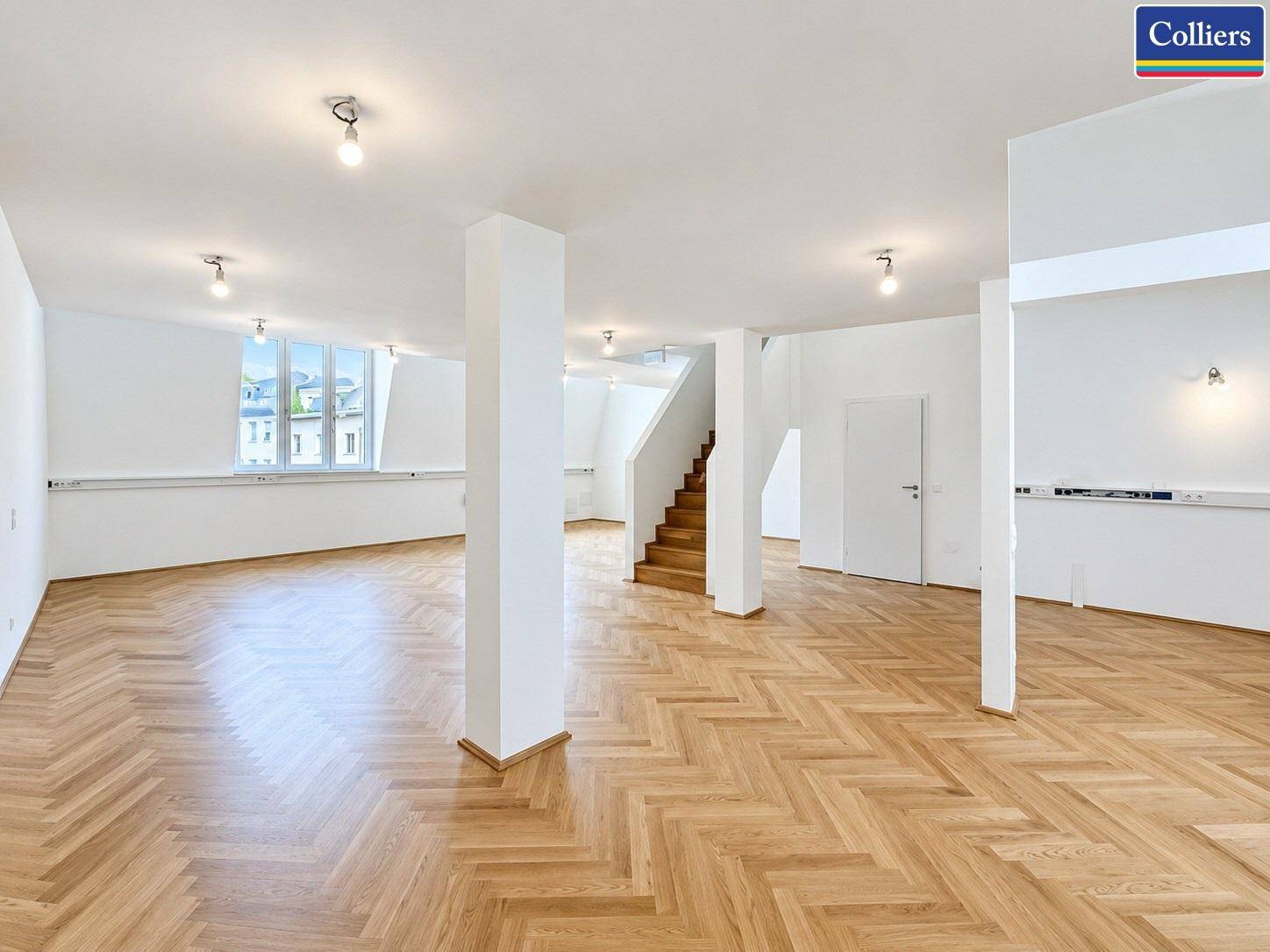 Weitläufiger offener Bereich mit Fischgrätparkett, weißen Säulen und Blick auf einen Treppenaufgang und Türen.