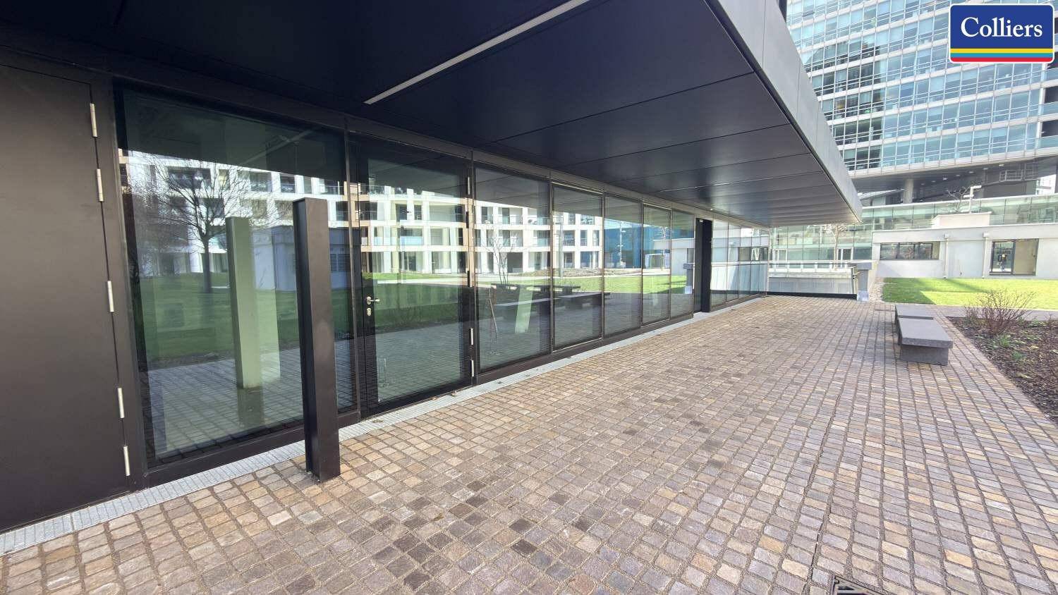 Moderne Gebäudefassade mit großen Glasflächen und einem gepflasterten Bereich davor, der zum Verweilen einlädt.