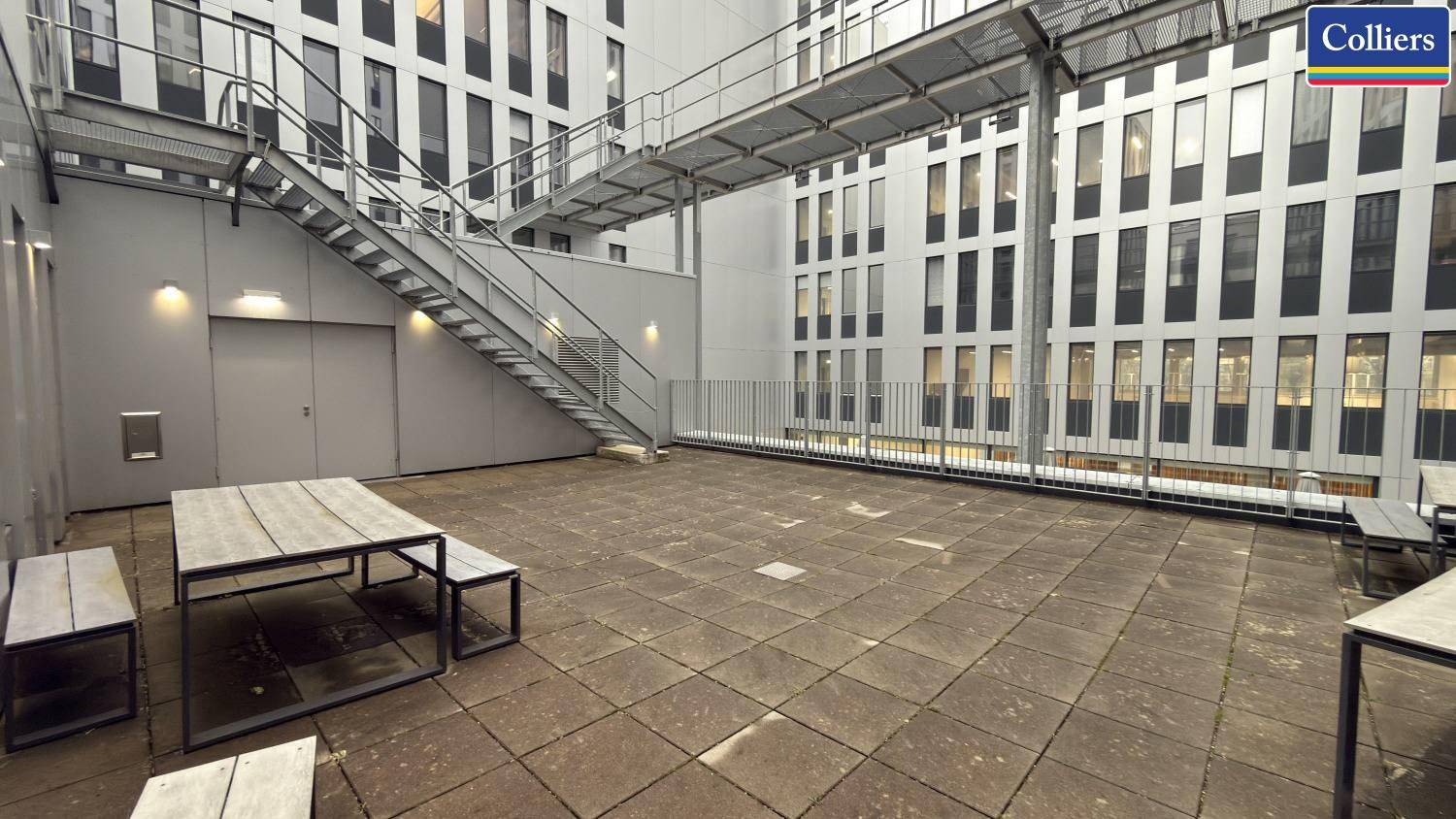 Große Außenterrasse mit Steinfliesen, Holztischen und Metalltreppe zwischen Gebäuden.