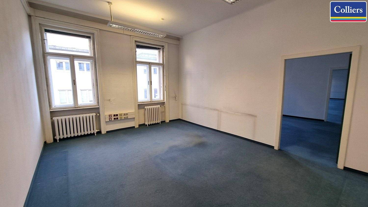 Helles Büro mit blauem Teppichboden, zwei Fenstern und Zugang zu einem weiteren Raum.