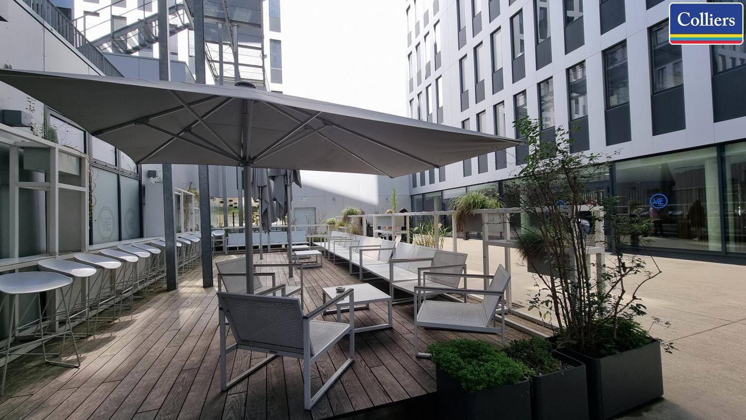 Außenbereich mit Holzterrasse, Sonnenschirm und modernen Sitzgelegenheiten, umgeben von Gebäuden.