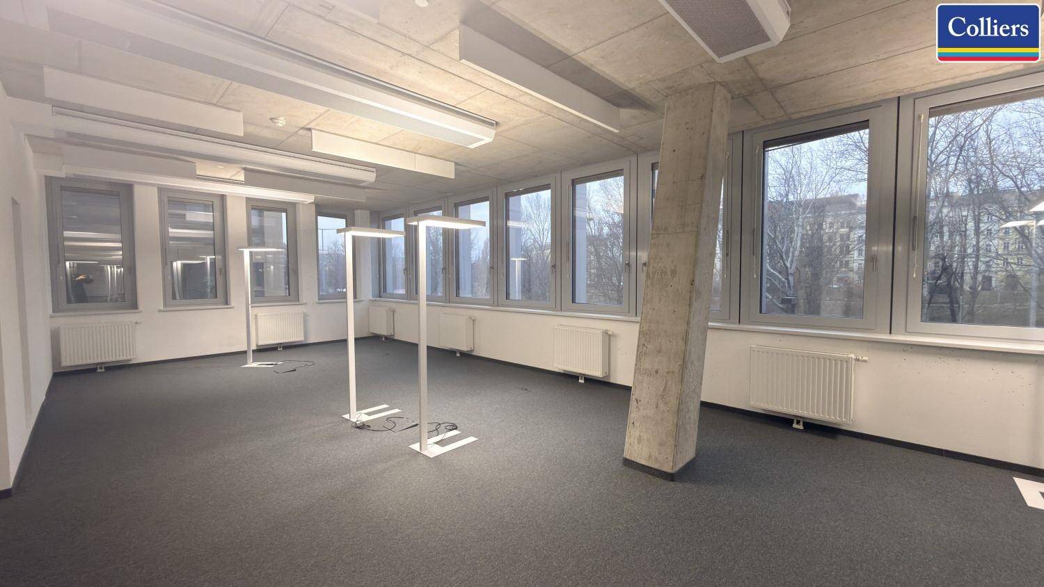 Unmöbliertes Büro mit Teppichboden, großen Fenstern und einem markanten Betonpfeiler in der Mitte des Raumes.