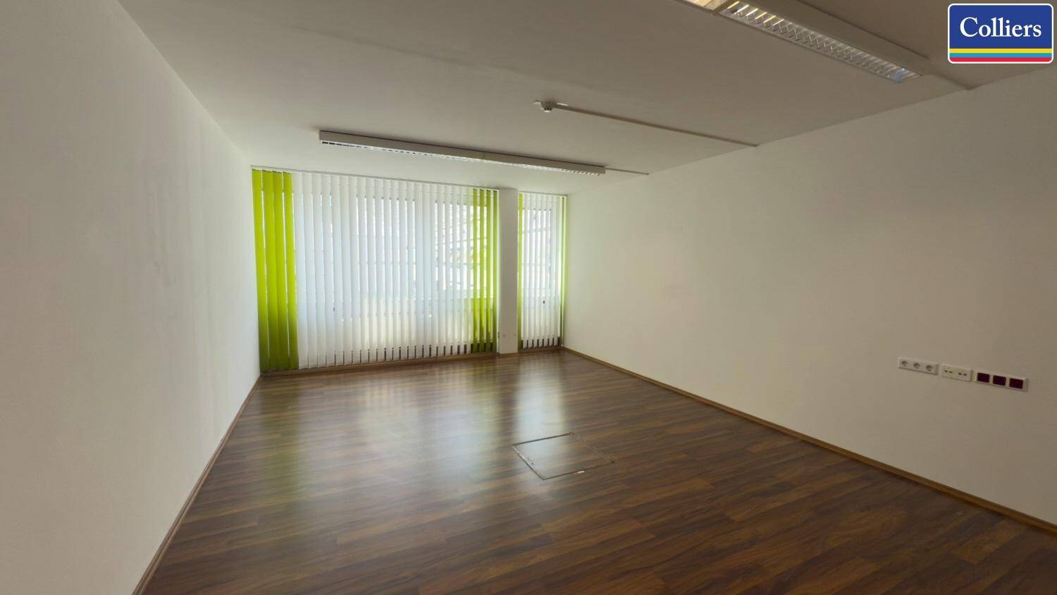 Helles Büro mit Holzboden und einer großen Fensterfront, die mit vertikalen Jalousien versehen ist.
