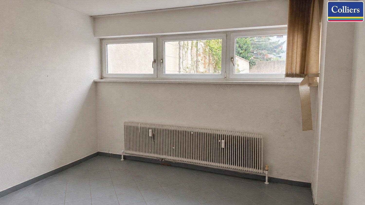 Kleiner Raum mit grauen Fliesen, einem Fenster und einem Heizkörper, der als Abstellraum dienen könnte.