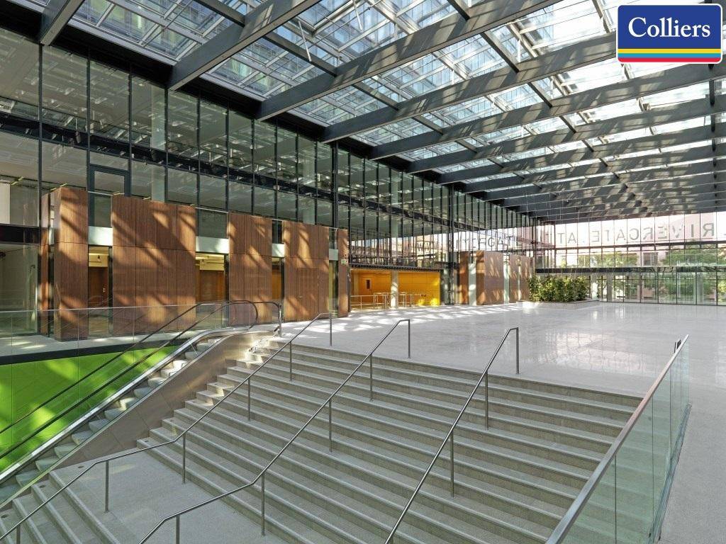 Geräumige, lichtdurchflutete Lobby mit hoher Glasdecke und breiter Treppe, die ein modernes Ambiente schafft.