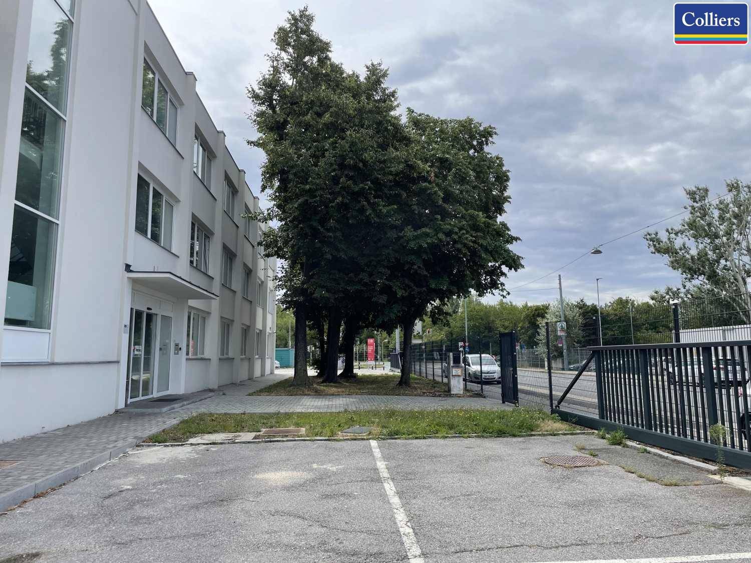 Außenansicht des Gebäudes mit Parkplätzen und Zufahrt zur Straße, umgeben von Bäumen.