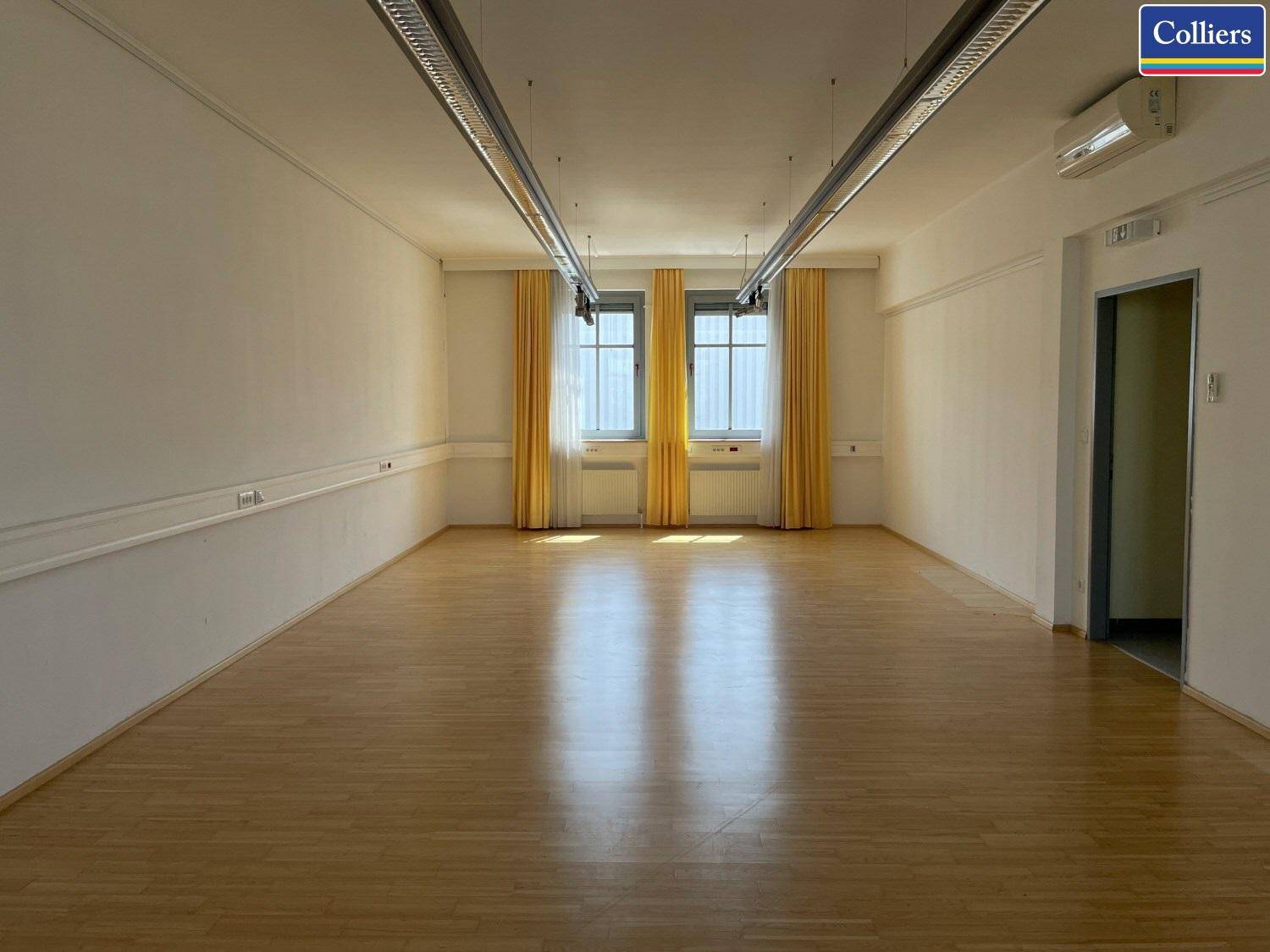 Langer, leerer Raum mit Holzfußboden, großen Fenstern und gelben Vorhängen, gut beleuchtet.