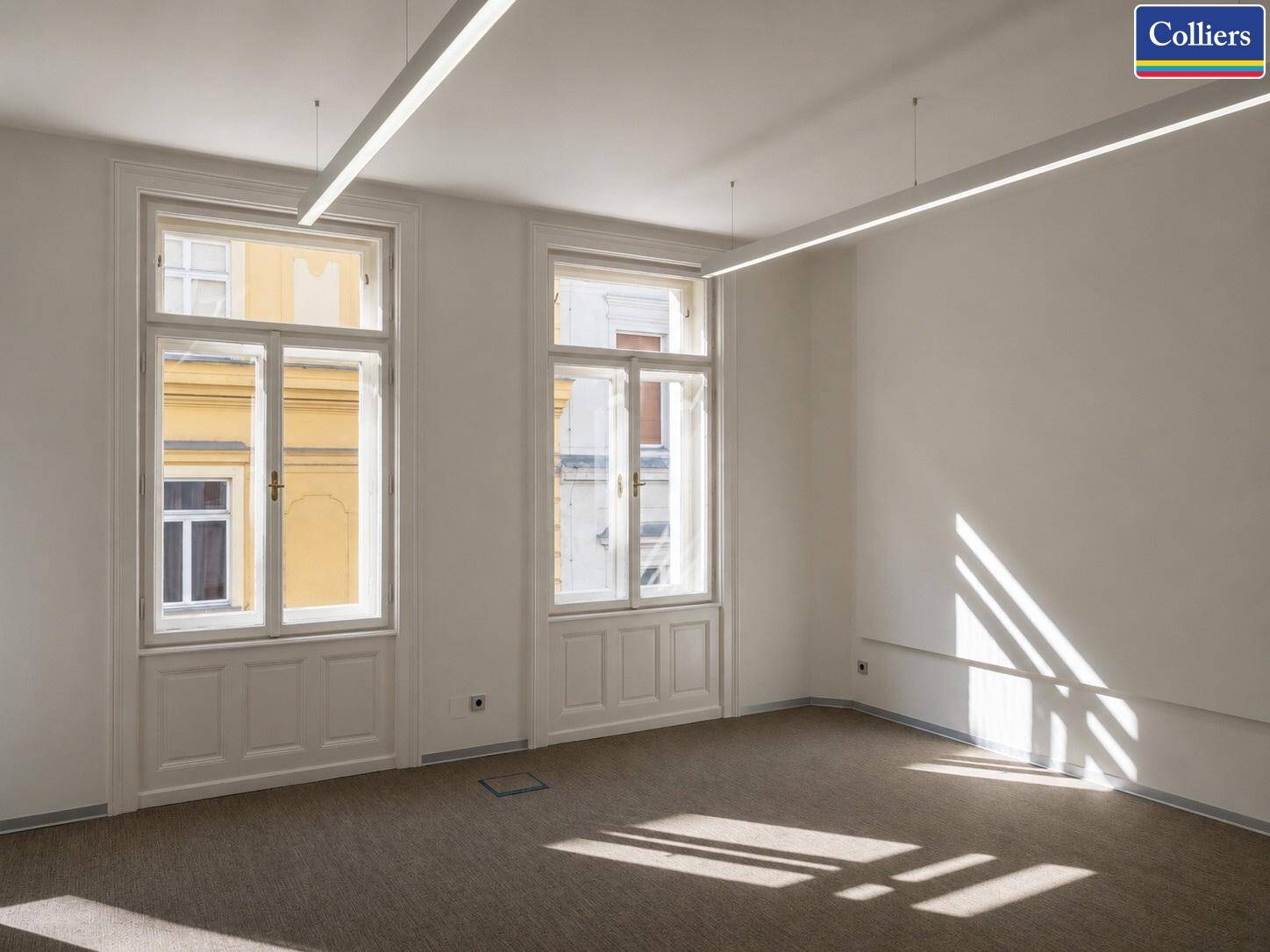 Großer, heller Büroraum mit zwei hohen Fenstern, Teppichboden und moderner Deckenbeleuchtung.