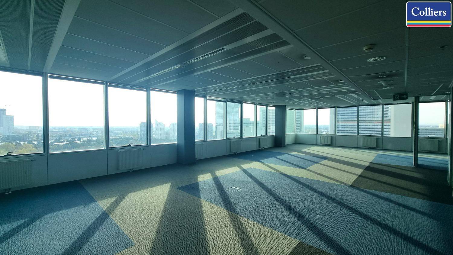 Helles Büro mit gemustertem Teppichboden und durchgehender Fensterfront, die viel Tageslicht hereinlässt.