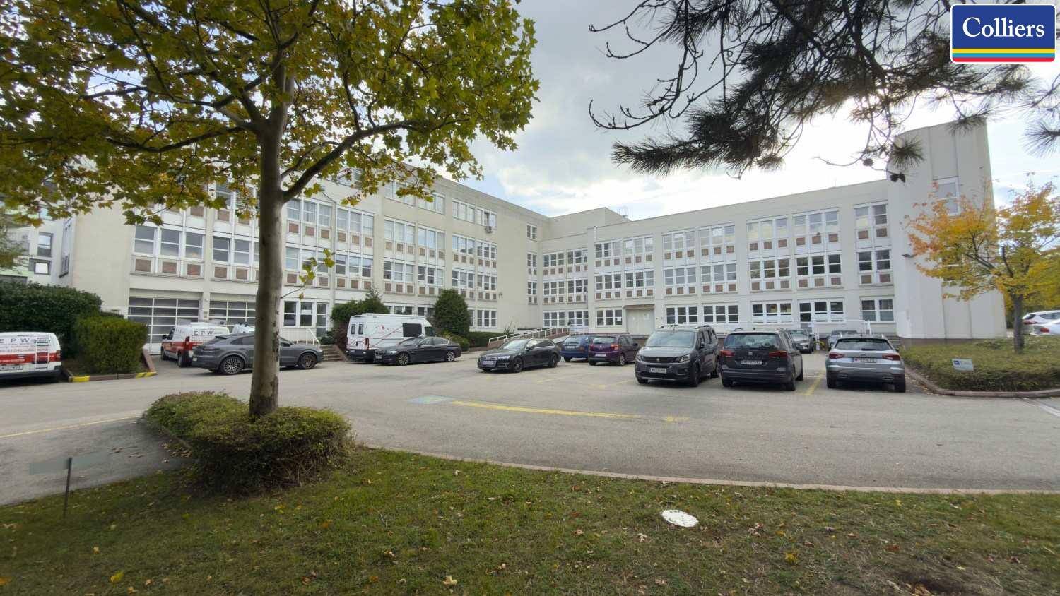 Großes Bürogebäude mit weitläufigem Parkplatz und Bäumen, umgeben von einer grünen Landschaft.