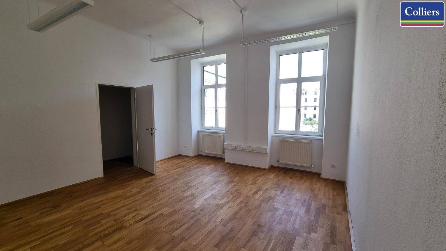 Ein geräumiger Büroraum mit Holzboden, weißen Wänden und einer offenen Tür, die zu einem weiteren Bereich führt.