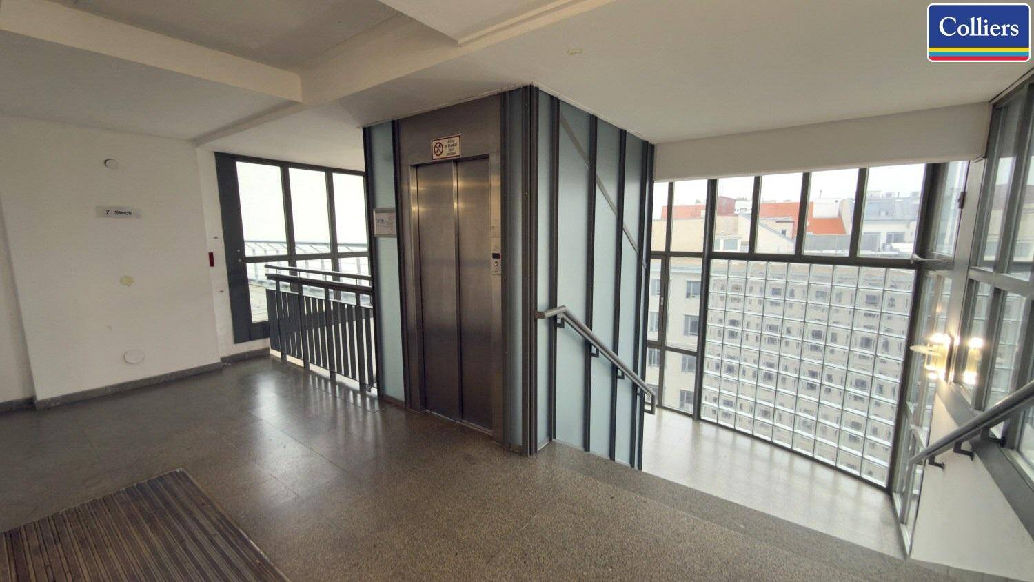 Heller Eingangsbereich mit Aufzug, Treppe und großen Fenstern, die viel Tageslicht hereinlassen.