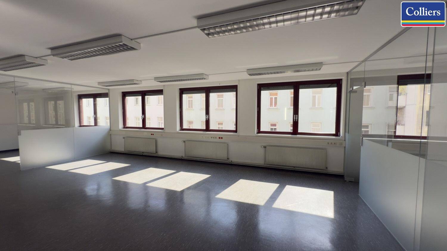 Helles Büro mit mehreren Fenstern, die viel Sonnenlicht hereinlassen, und Blick auf umliegende Gebäude.