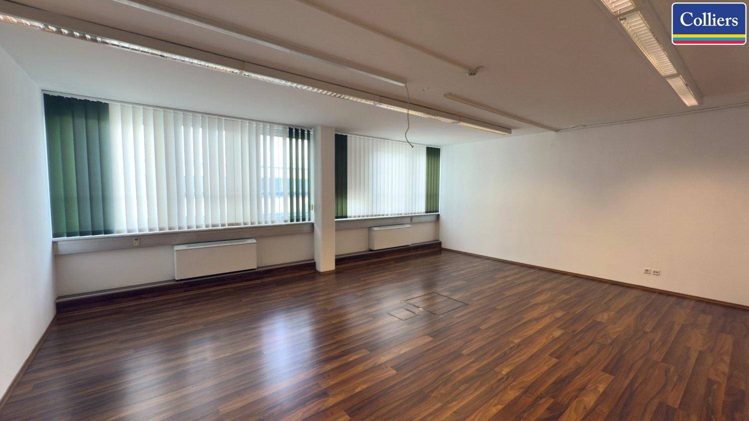 Geräumiges Büro mit Holzboden und mehreren Fenstern, die mit vertikalen Jalousien ausgestattet sind.