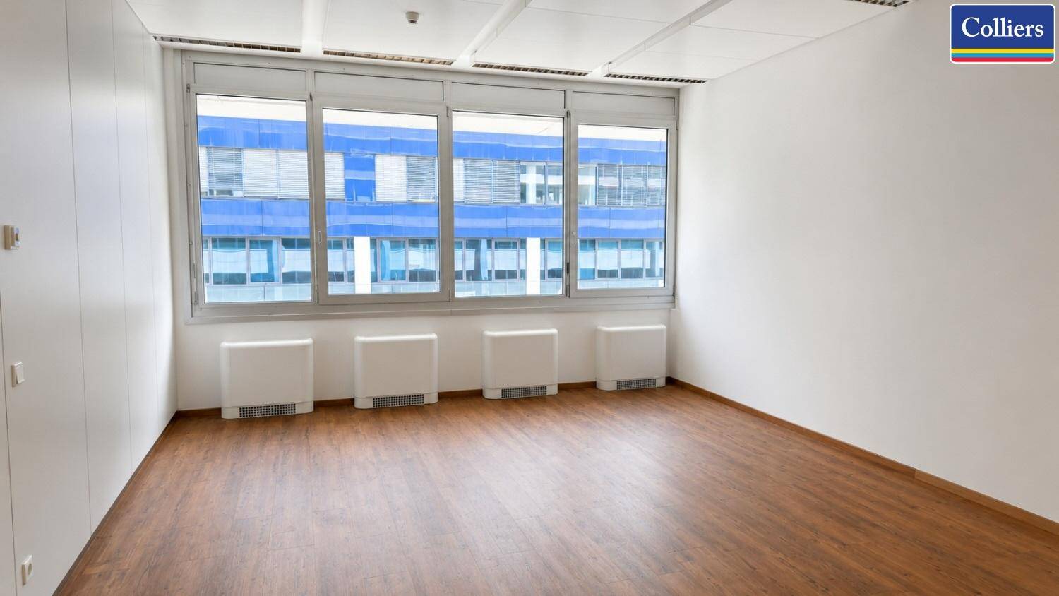 Helles Büro mit Holzboden, großen Fenstern und Blick auf ein gegenüberliegendes Gebäude.
