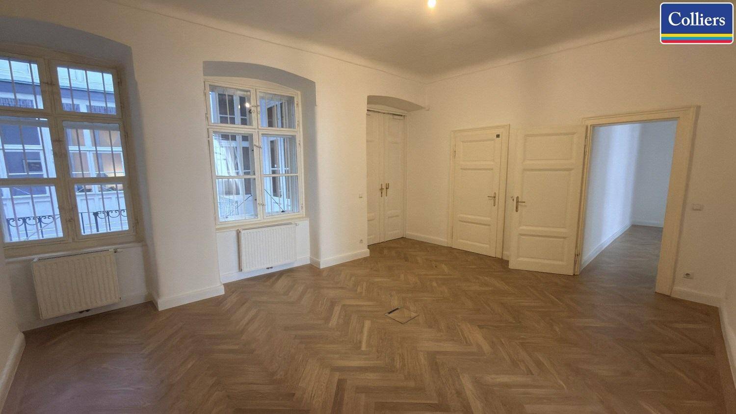 Großer Raum mit Fischgrätparkett, hohen Fenstern und mehreren Türen, ideal für flexible Büronutzung.