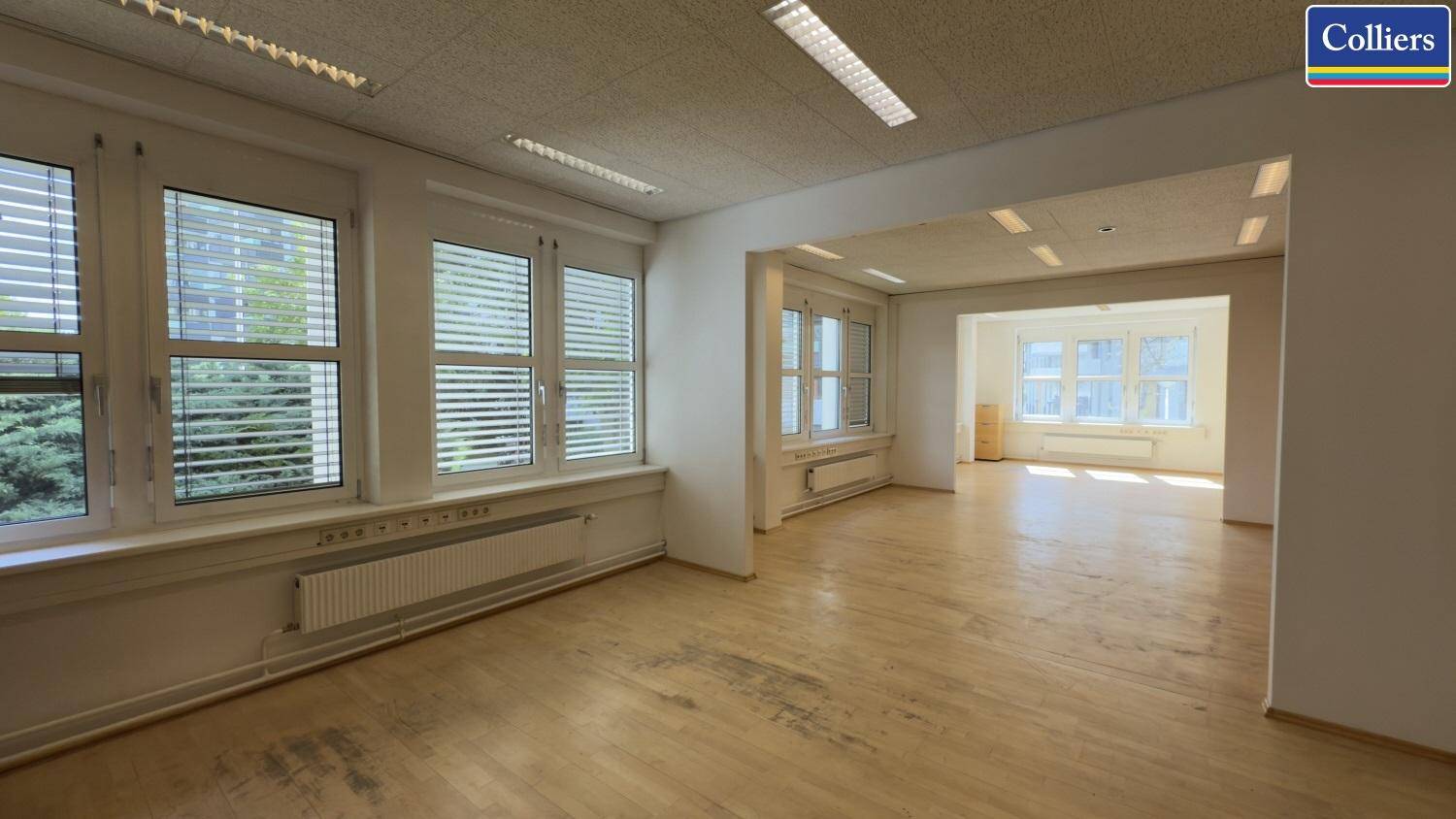 Offener Bürobereich mit durchgehenden Fenstern und hellem Parkettboden, der in einen weiteren Raum führt.