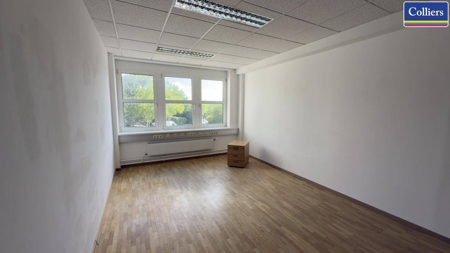 Leeres Büro mit hellem Holzfußboden, weißen Wänden und einem großen Fenster, das viel Tageslicht hereinlässt.