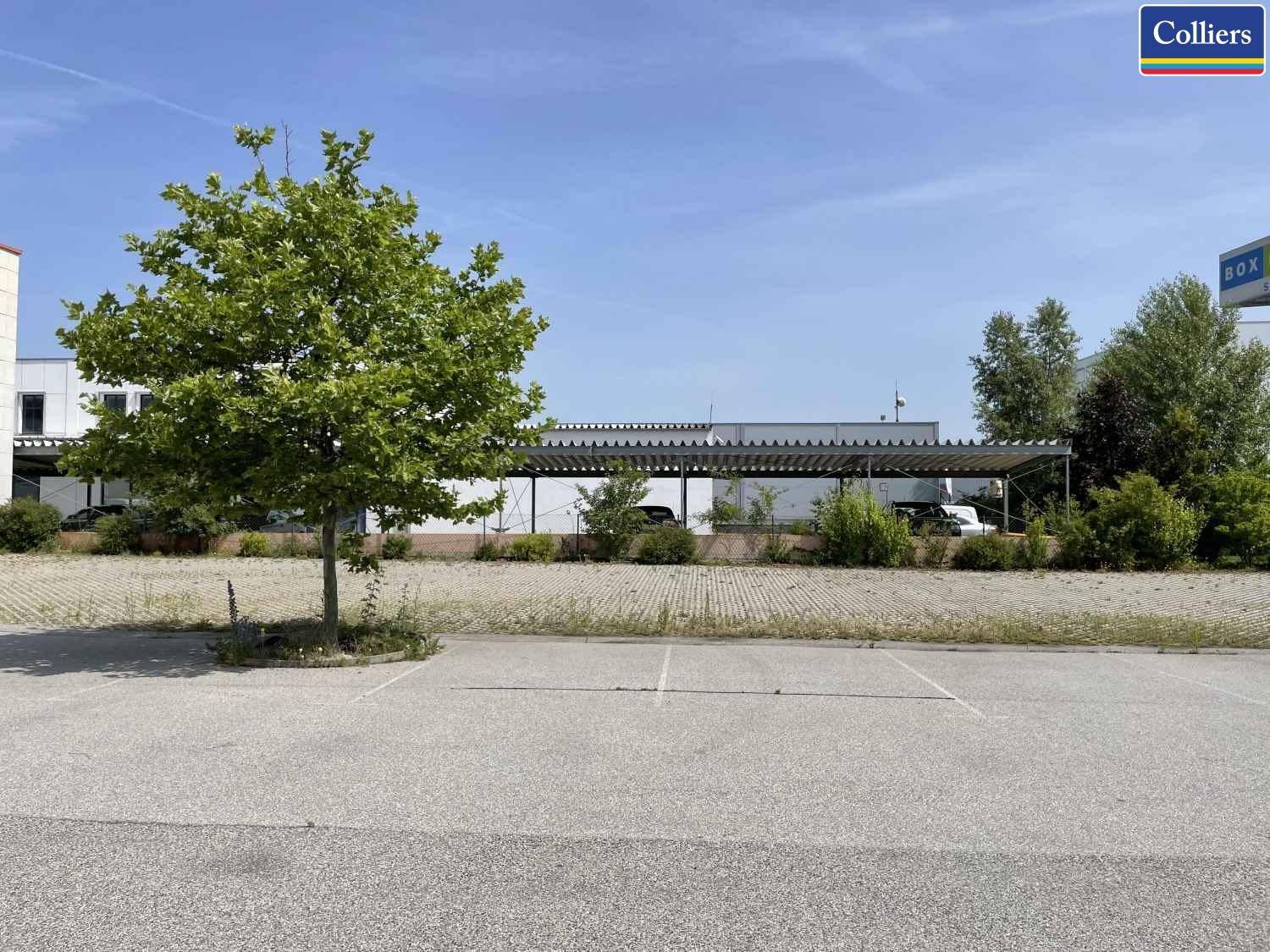 Großer Parkplatz mit einem Baum und überdachten Stellplätzen im Hintergrund unter blauem Himmel.
