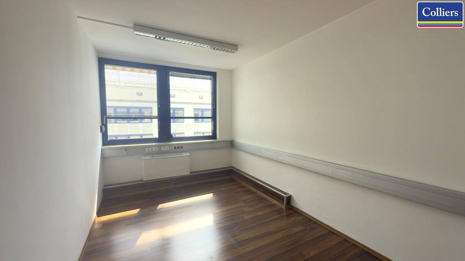 Bürofläche mit Holzboden und einem Fenster, das Tageslicht hereinlässt und einen Blick auf ein Gebäude ermöglicht.