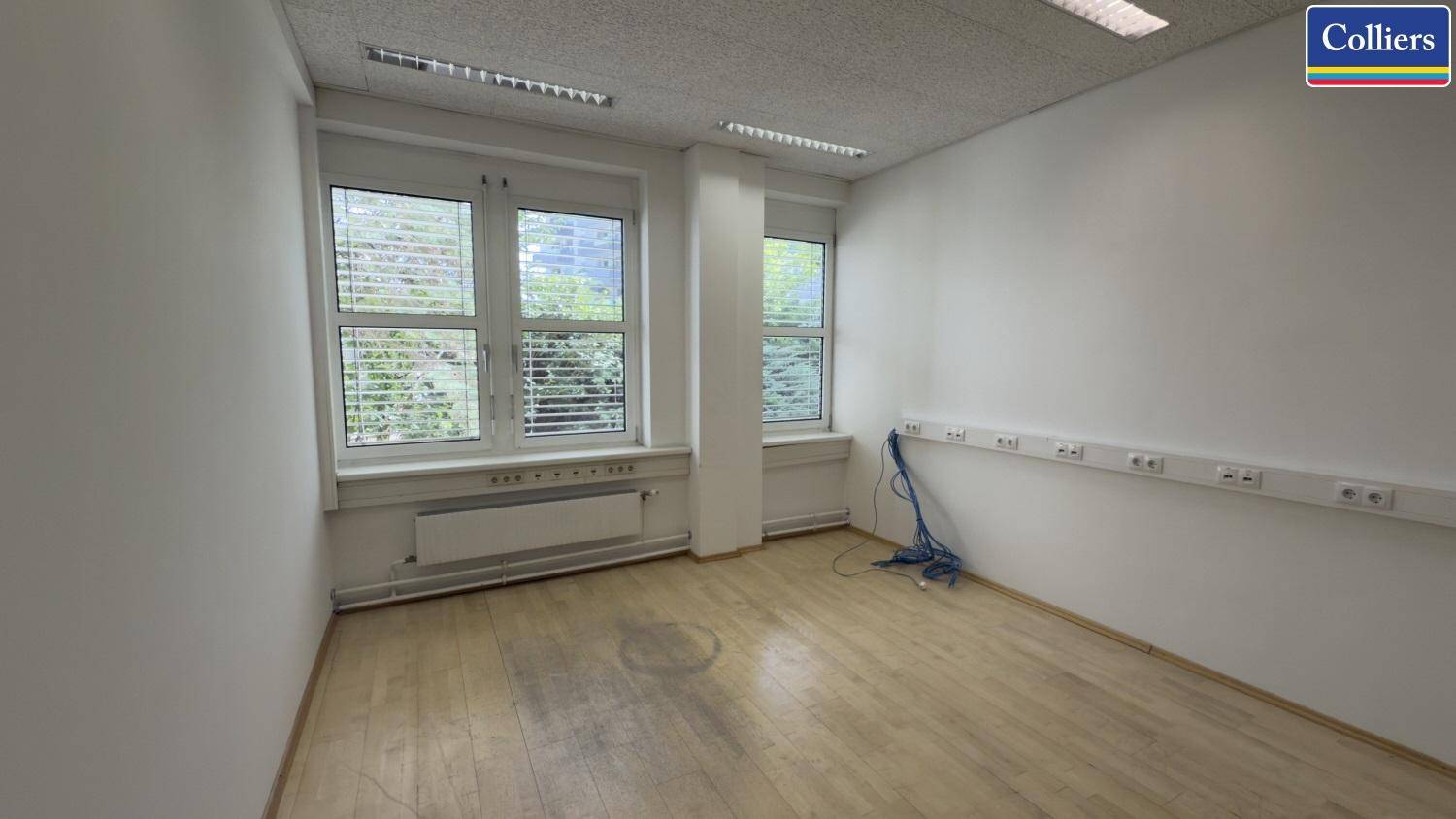 Leeres Büro mit hellem Holzfußboden, weißen Wänden und großen Fenstern mit Jalousien.
