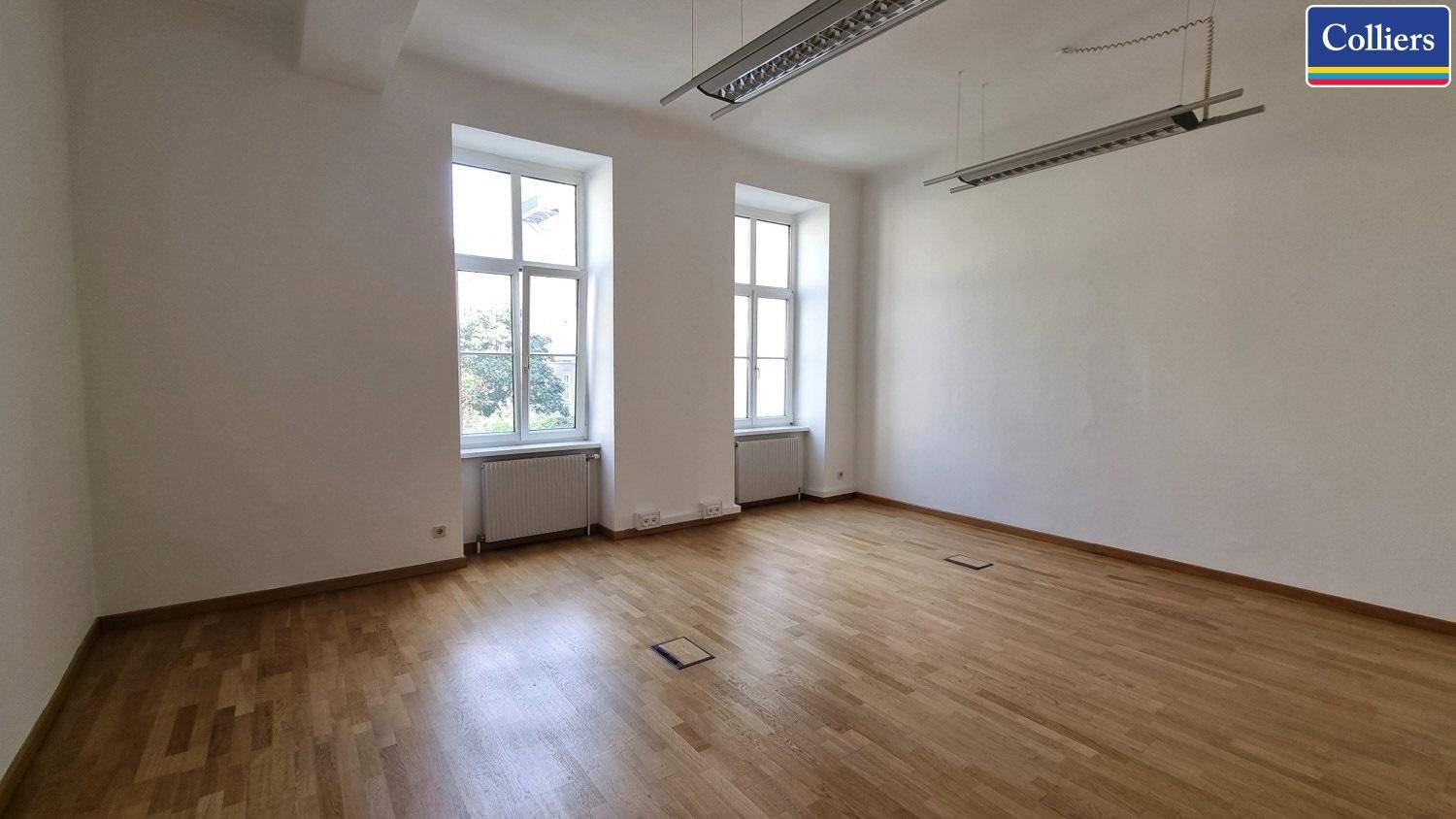 Ein heller, leerer Büroraum mit Holzboden und zwei großen Fenstern, die viel Tageslicht hereinlassen.