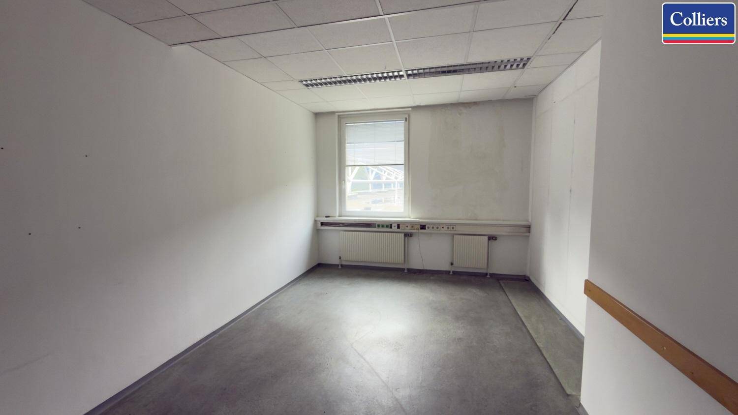 Einfacher Büroraum mit Betonboden, einem Fenster und schlichter Wandgestaltung, der renoviert werden muss.