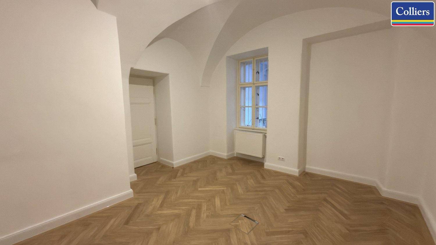 Raum mit gewölbter Decke und Fischgrätparkett, ausgestattet mit einem Fenster und Heizkörper in einem Altbau.
