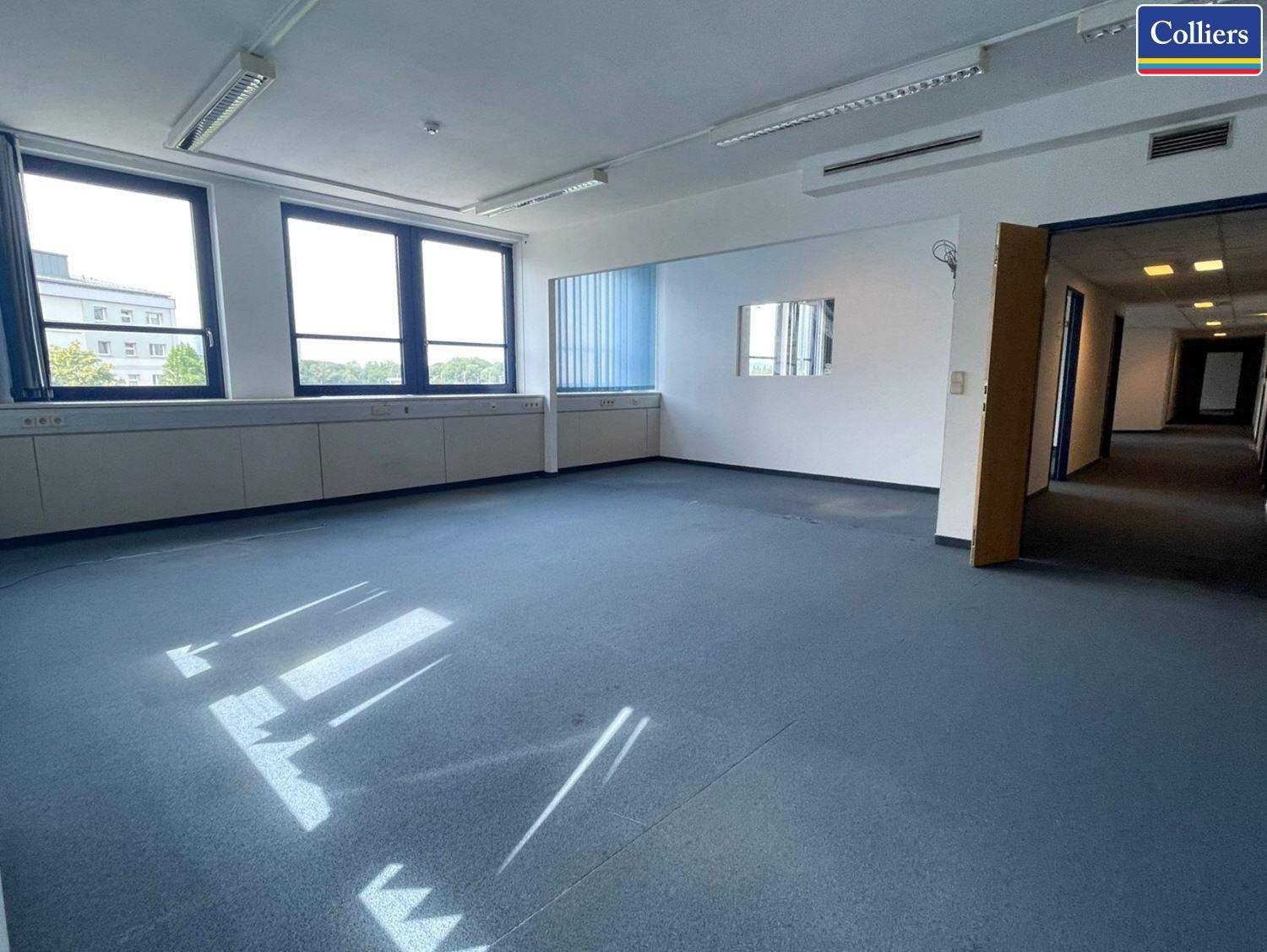Geräumiges leeres Büro mit Teppichboden, großen Fenstern und Zugang zu einem Flur, ideal für flexible Nutzung.