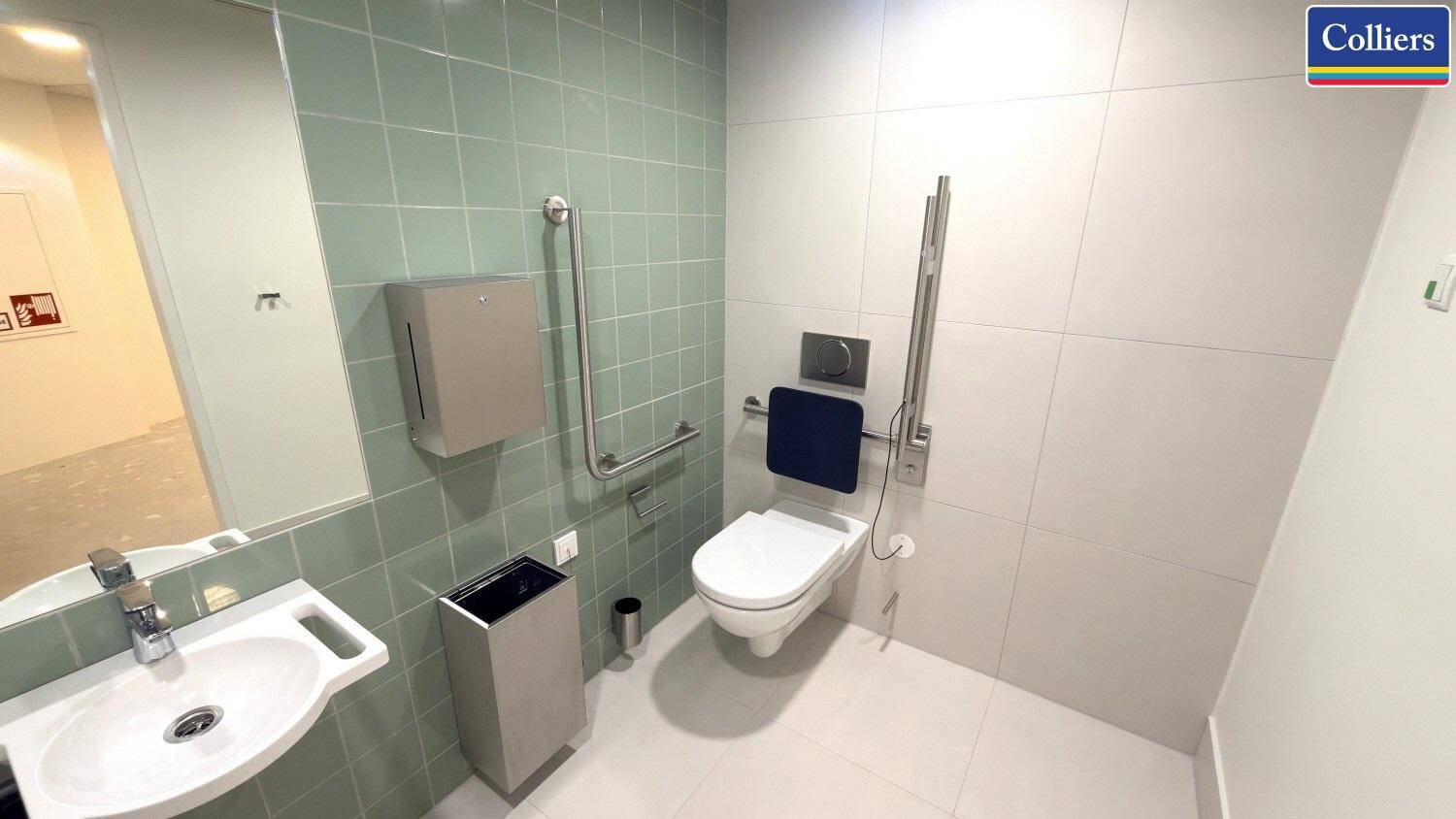 Barrierefreies WC mit Haltegriffen, wandhängender Toilette und modernem Waschbecken, gefliest in Grün und Weiß.