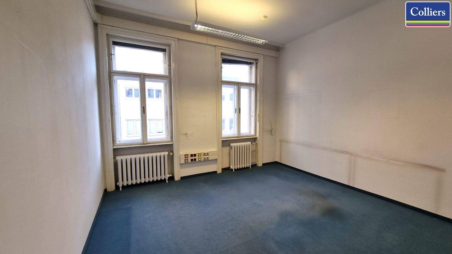 Leeres Büro mit blauem Teppichboden, Heizkörpern unter den Fenstern und Deckenleuchte.
