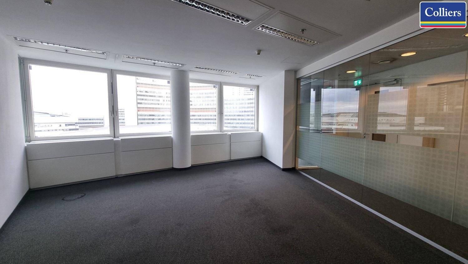 Heller Büroraum mit Teppichboden, großen Fenstern und einer Glaswand zu einem angrenzenden Bereich, ideal für konzentriertes Arbeiten.